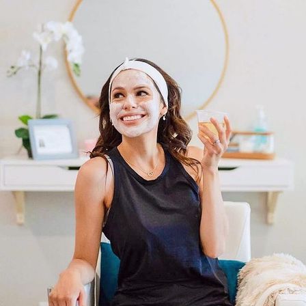 Smiling woman in a headband with a white facial mask, relaxing with a drink at a bright home vanity — at-home spa and skincare moment.