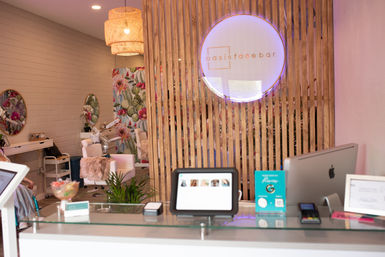 Bright modern spa and nail salon reception with glass counter, tablet and iMac, wooden slat divider with glowing round sign, floral wallpaper and plush pedicure chairs.