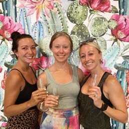Three smiling friends in casual summer outfits holding champagne flutes in front of a colorful cactus-and-flower mural backdrop, cheerful celebration vibe.