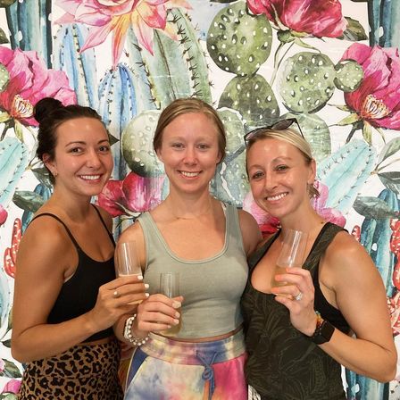 Three smiling friends in casual summer outfits holding champagne flutes in front of a colorful cactus-and-flower mural backdrop, cheerful celebration vibe.