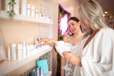 Two women browsing skincare products on white shelves in a bright boutique spa; one points to a bottle while the other holds a wine glass, neon sign and plants in the background.