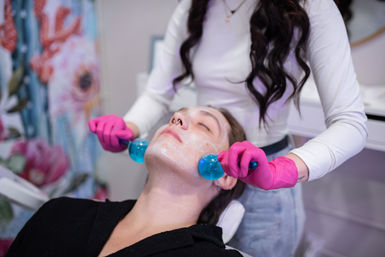 Spa facial treatment: esthetician wearing pink gloves uses blue cooling globes to massage cream-covered cheeks of a relaxed client in a floral treatment room.
