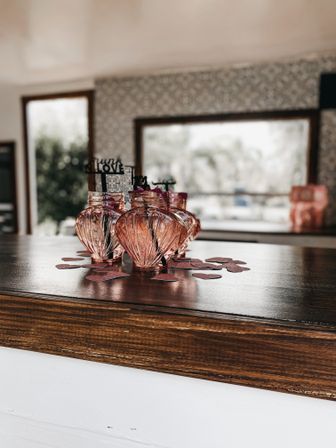 Pink glass jar centerpiece on a wooden kitchen island with scattered heart confetti and 'LOVE' picks, sunlit window and patterned wallpaper blurred in the background