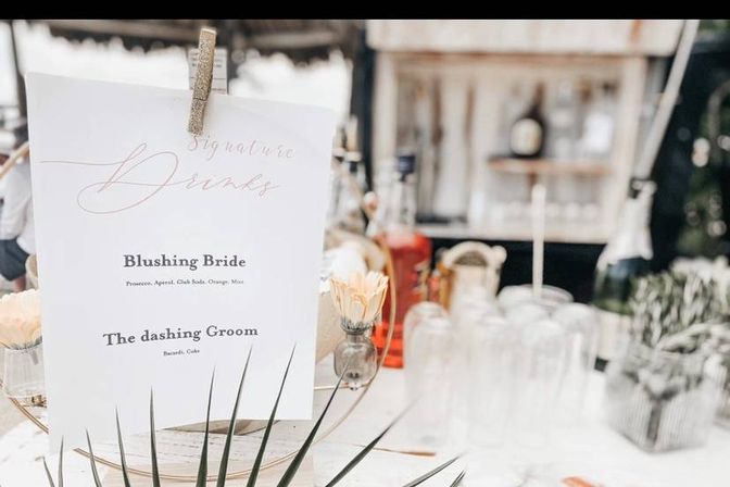 Rustic wedding bar with a 'Signature Drinks' menu listing 'Blushing Bride' and 'The dashing Groom', surrounded by bottles, stacked glassware, small floral accents, and tropical greenery.