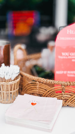 Close-up tabletop scene with stacked pale-pink napkins printed with a small red heart, a woven wicker basket and a rattan cup holding white cocktail picks against a soft, blurred background.