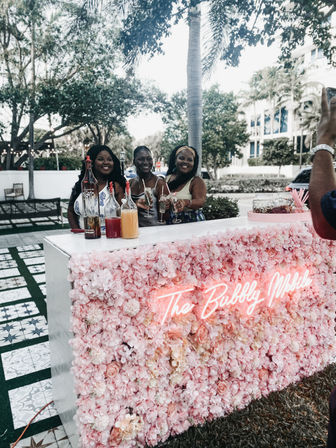 Smiling trio toasting at a pink flower‑covered pop-up mimosa bar outdoors under palm trees, bottles and juice pitchers on the counter and a neon sign glowing