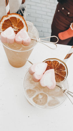 Top-down view of two iced citrus cocktails in clear plastic cups, each garnished with a dried orange wheel, skewered pink heart-shaped marshmallows and a striped paper straw on a white table — refreshing summer drink shot.