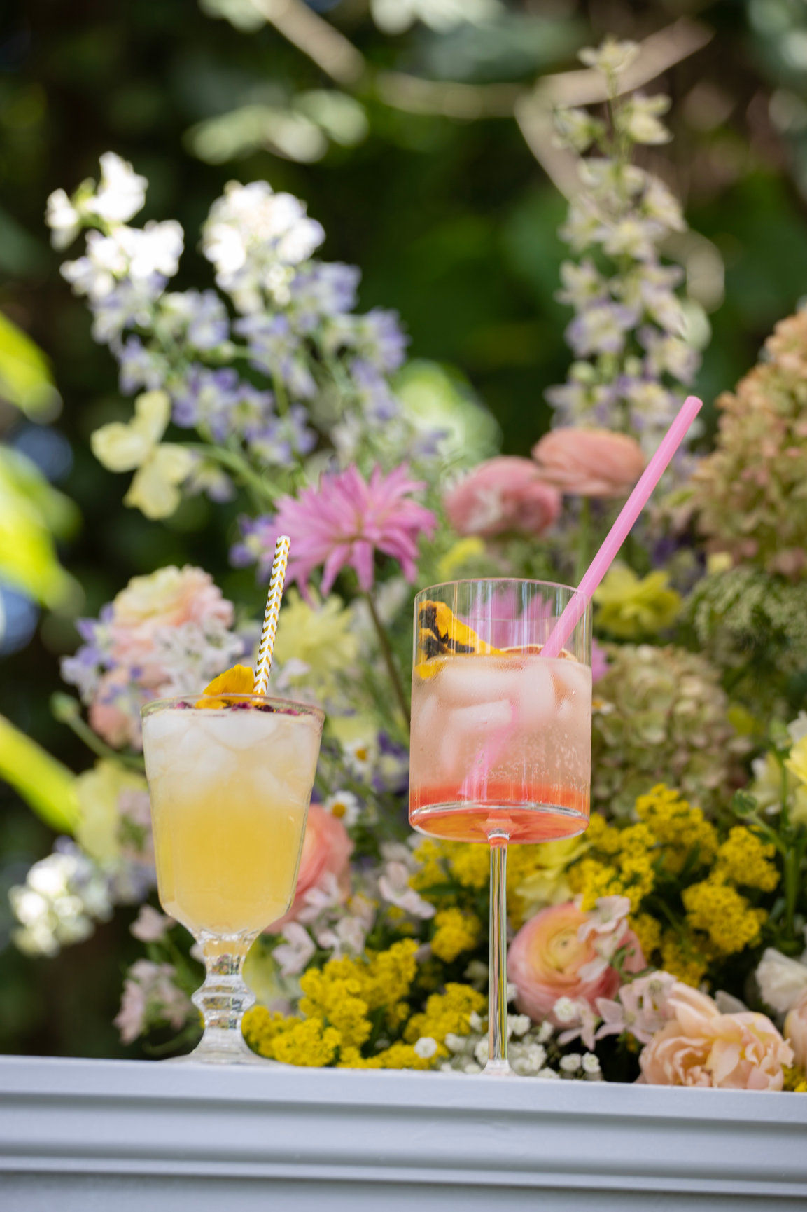 Two iced summer cocktails — a yellow drink and a pink spritz with straws and edible flower garnishes — set on a white ledge against a lush garden floral backdrop