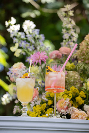 Two iced summer cocktails — a yellow drink and a pink spritz with straws and edible flower garnishes — set on a white ledge against a lush garden floral backdrop