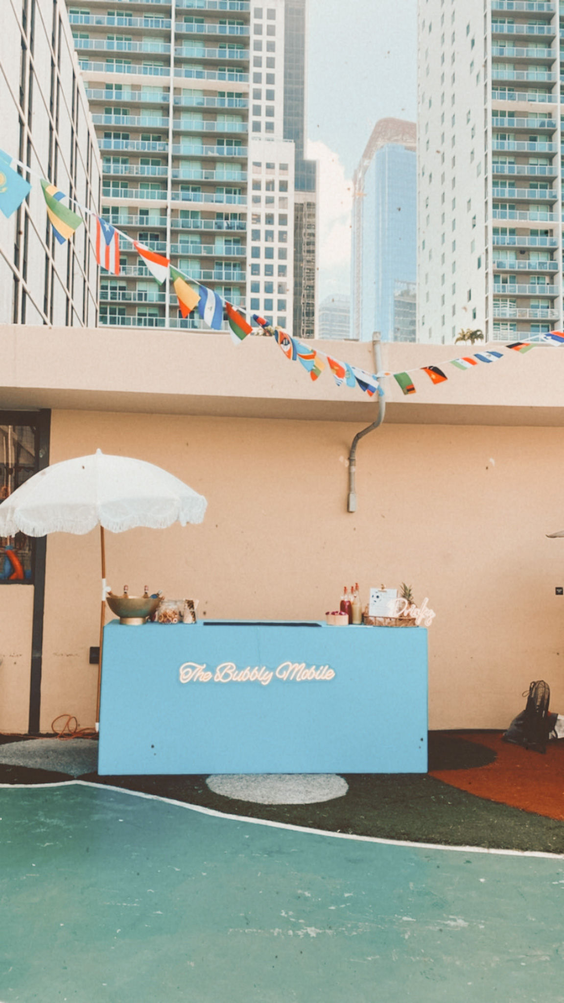 Pastel blue pop-up drink cart with a white umbrella on a sunny urban rooftop terrace, colorful pennant flags overhead and downtown high-rise buildings in the background.