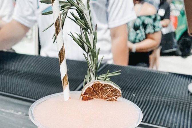 Pink frozen cocktail in a clear plastic cup with a striped paper straw, rosemary sprig and dried citrus wheel at an outdoor summer bar