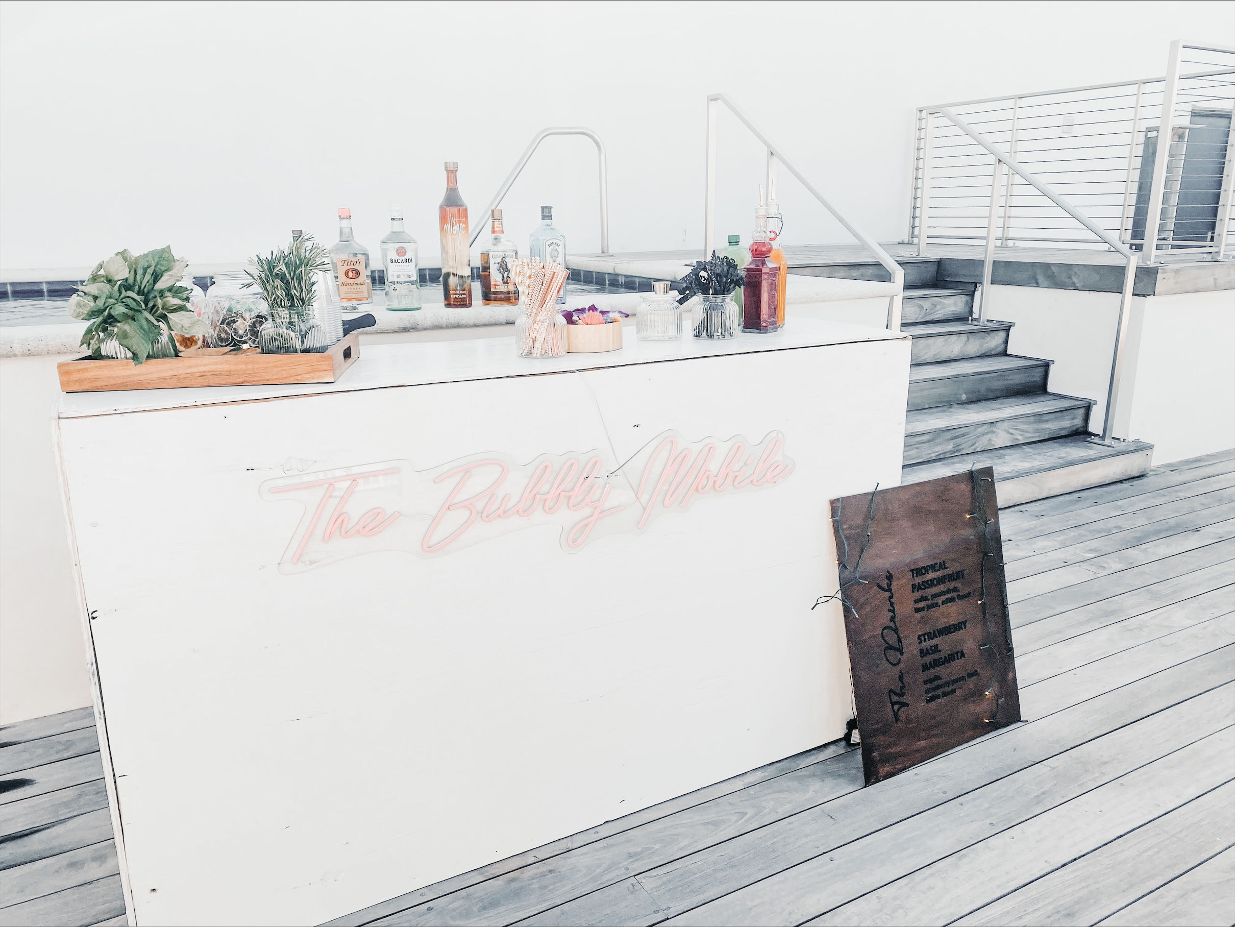 Rooftop poolside cocktail bar on a light wooden deck — white serving counter with bottles, garnishes, potted herbs, neon script sign, and metal stairs and railings.