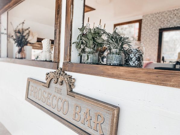 Rustic white-and-wood bar counter with an ornate carved sign, glass jars of fresh herbs and straws, and a bright airy café interior.