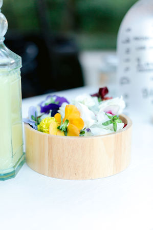 Spring garden table scene: round wooden bowl filled with colorful edible flowers (pansies and violas) beside a glass bottle of pale green drink on a white surface