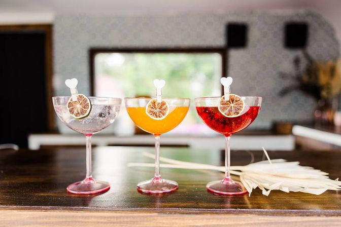 Three colorful craft cocktails—clear, orange, and red—served in coupe glasses on a wooden bar counter, each garnished with a dried citrus wheel clipped by a tiny heart pick, blurred modern bar interior in the background.