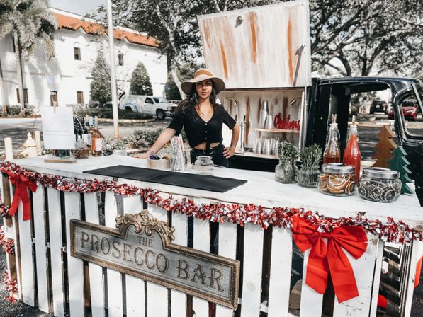 Outdoor mobile prosecco bar at a sunny street market, woman in a sun hat behind a white wooden counter decorated with red bows and garlands, bottles, jars of garnishes and greenery on display