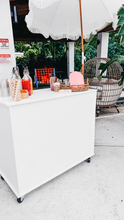 White rolling refreshment cart under a fringed umbrella on a tropical patio, with glass juice bottles, paper straws, wicker basket, cups, and a nearby rattan hanging chair