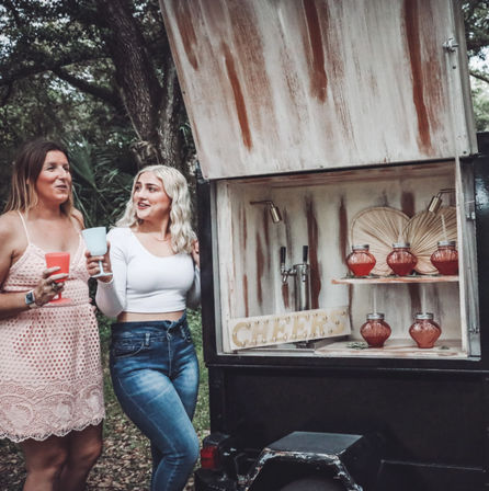 Two friends holding colorful cups at an outdoor wooded backyard party beside a rustic mobile bar trailer with taps, mason-jar cocktails on shelves and a “CHEERS” sign.