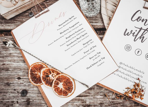 Cocktail drink menu on clipboards with dried citrus slices, a twisted bar spoon and glass on a rustic wooden table