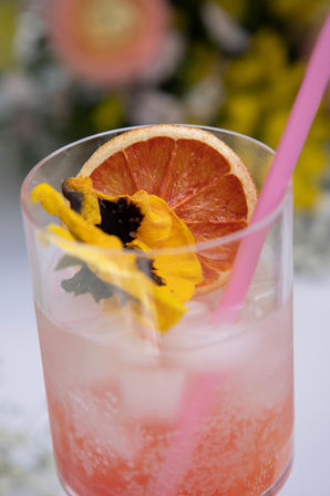 Close-up of a pink sparkling grapefruit cocktail with ice, dried grapefruit wheel, yellow edible pansy garnish and pink straw — a refreshing summer drink