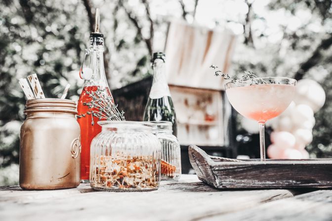 Outdoor rustic wooden table with a pink coupe cocktail garnished with thyme, mason jar with paper straws, glass bottles and jars of dried flowers in a garden party setting with blurred balloons