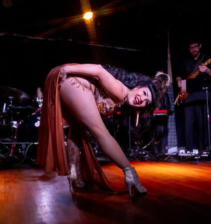 Burlesque dancer in a feathered headpiece and sequined costume bends forward on a cabaret stage, fishnet stockings and studded heels glinting under warm spotlights with a live band in the background.