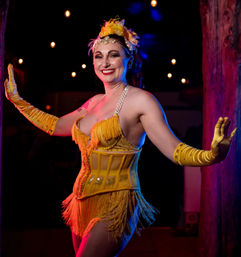 Smiling burlesque performer in a yellow fringed corset, long gloves and floral headpiece on a dim cabaret stage with warm string lights