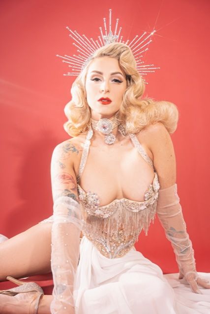 Glam vintage Hollywood-style burlesque performer seated against a red backdrop, wearing a sparkling halo crown, platinum blonde finger-wave hair, red lipstick, beaded fringe corset, sheer gloves, visible arm tattoos and a flowing white skirt.
