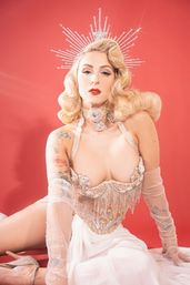 Glam vintage Hollywood-style burlesque performer seated against a red backdrop, wearing a sparkling halo crown, platinum blonde finger-wave hair, red lipstick, beaded fringe corset, sheer gloves, visible arm tattoos and a flowing white skirt.