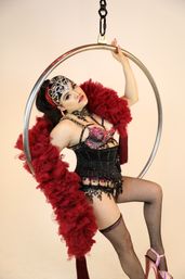 Burlesque performer in jeweled corset, ornate headpiece and red feather boa posing on a suspended aerial hoop in a studio, fishnet stockings and pink platform heels.