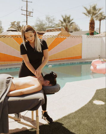 Relaxing poolside massage in a sunny backyard: therapist giving a back massage on a portable table beside a swimming pool, colorful geometric mural, palm trees, and a pink inflatable float.