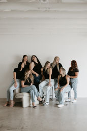 Eight women in a bright minimalist studio wearing black tops and light denim, laughing and chatting while seated on chairs and a low couch against a white wall — casual group portrait.