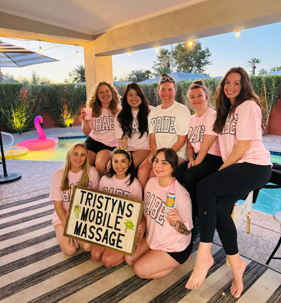 Group of eight women at a backyard poolside bachelorette party wearing matching pink tees and a white "BRIDE" shirt, holding a wooden sign promoting mobile massage with an inflatable flamingo, patio string lights, and palm trees at sunset.