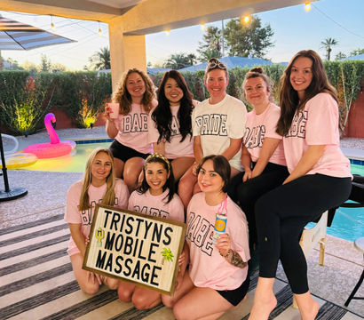 Group of eight women at a backyard poolside bachelorette party wearing matching pink tees and a white "BRIDE" shirt, holding a wooden sign promoting mobile massage with an inflatable flamingo, patio string lights, and palm trees at sunset.