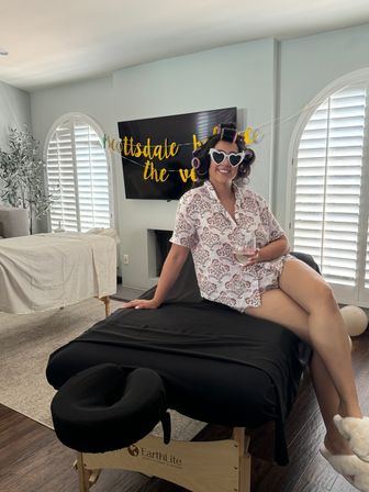 Person in floral pajamas with hair rollers and heart-shaped sunglasses sitting on a black-covered massage table holding a wine glass in a bright Scottsdale living room with arched shuttered windows and a gold party banner.