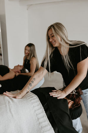 Blonde massage therapist with geometric forearm tattoo giving a back massage to a client in a bright wellness studio, another therapist working with a client in the background
