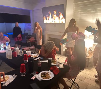 Evening backyard patio party with guests around a long black-covered table of plates, red cups and drinks, a wall-mounted TV displaying flames, a lit lamp and people playfully holding colorful toy water guns near a pool.