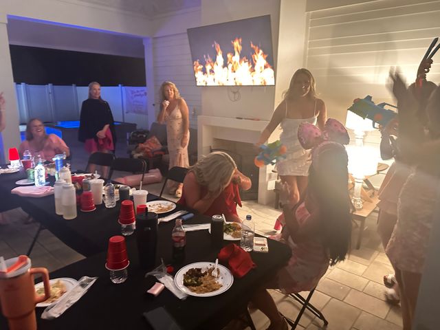 Evening backyard patio party with guests around a long black-covered table of plates, red cups and drinks, a wall-mounted TV displaying flames, a lit lamp and people playfully holding colorful toy water guns near a pool.