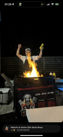 Nighttime outdoor hibachi chef performing a teppanyaki-style flame show on a portable grill cart, mustard bottle raised, condiments and coolers on a patio in Santa Rosa.