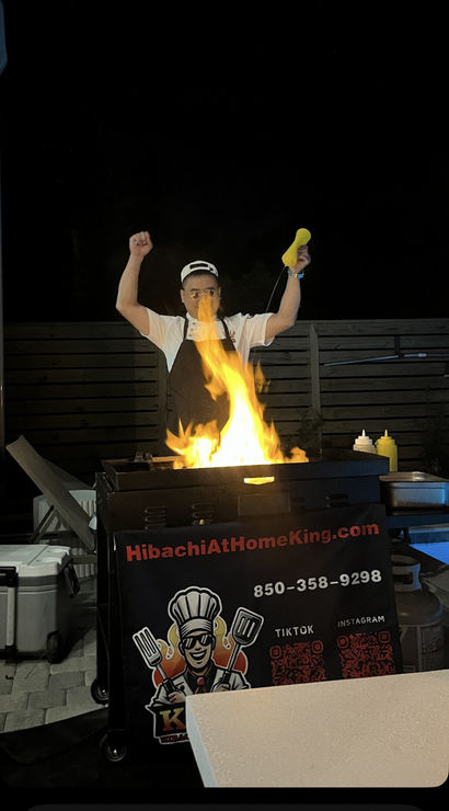 Nighttime outdoor hibachi chef performing a teppanyaki-style flame show on a portable grill cart, mustard bottle raised, condiments and coolers on a patio in Santa Rosa.