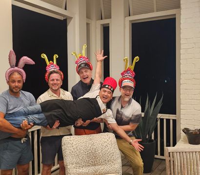 Five men on a covered porch at night wearing playful headgear (balloon bunny ears and colorful decorative hats) lifting a laughing friend horizontally — outdoor deck party with TV and fireplace in the background.