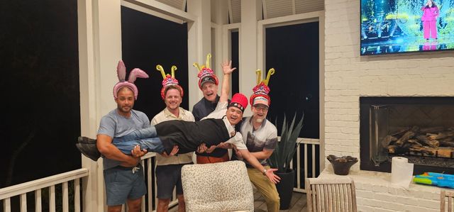 Five men on a covered porch at night wearing playful headgear (balloon bunny ears and colorful decorative hats) lifting a laughing friend horizontally — outdoor deck party with TV and fireplace in the background.