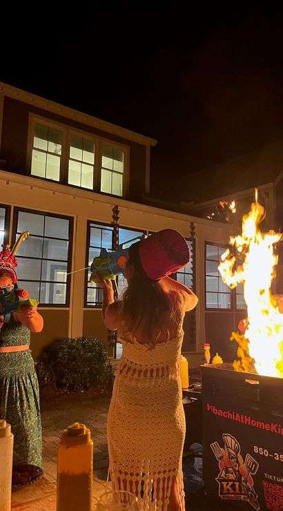 Nighttime backyard party by lit house windows, people in festive hats aiming colorful toy water guns at a large hibachi fire on an outdoor grill with condiment bottles in the foreground.