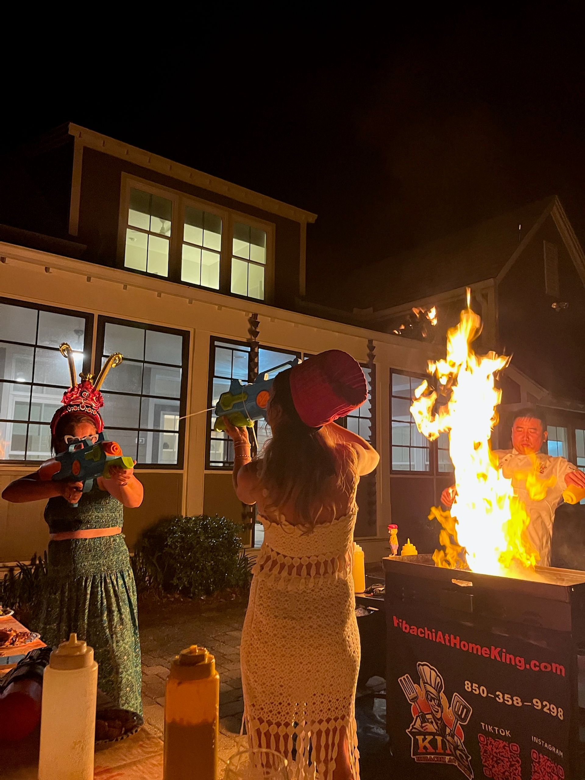 Nighttime backyard party by lit house windows, people in festive hats aiming colorful toy water guns at a large hibachi fire on an outdoor grill with condiment bottles in the foreground.