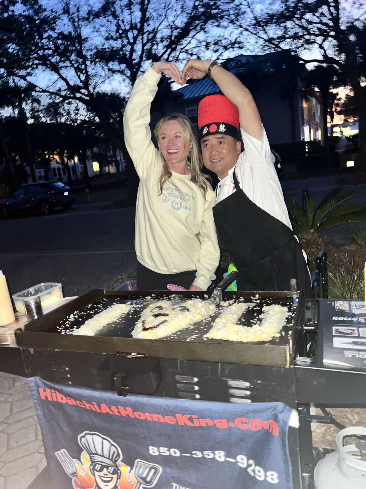 Smiling woman and hibachi chef in a red hat making a heart shape over an outdoor teppanyaki griddle at dusk, rice and sauce art visible on the hotplate.