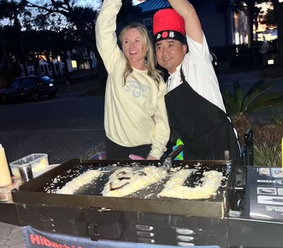 Smiling woman and hibachi chef in a red hat making a heart shape over an outdoor teppanyaki griddle at dusk, rice and sauce art visible on the hotplate.