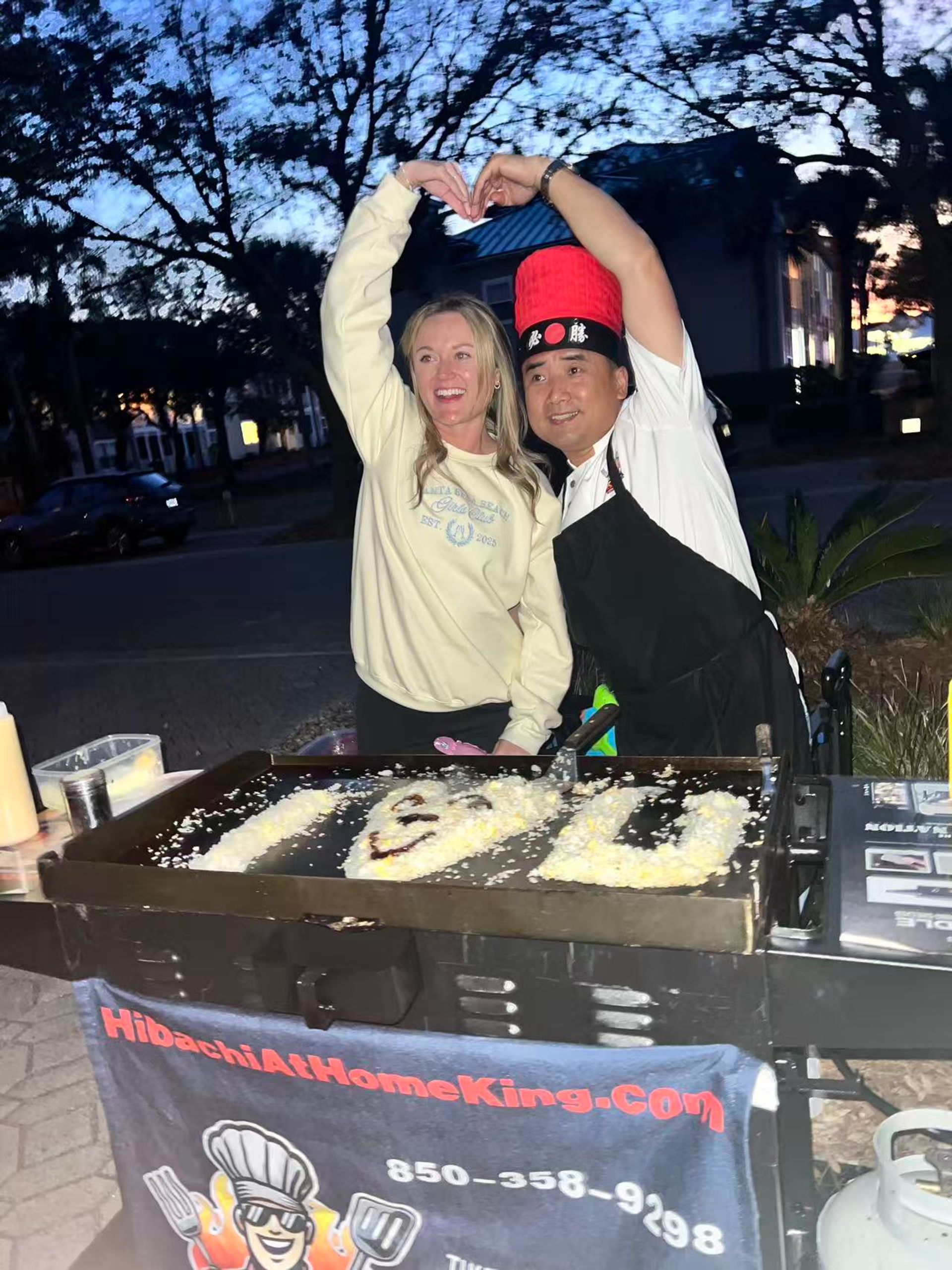 Smiling woman and hibachi chef in a red hat making a heart shape over an outdoor teppanyaki griddle at dusk, rice and sauce art visible on the hotplate.