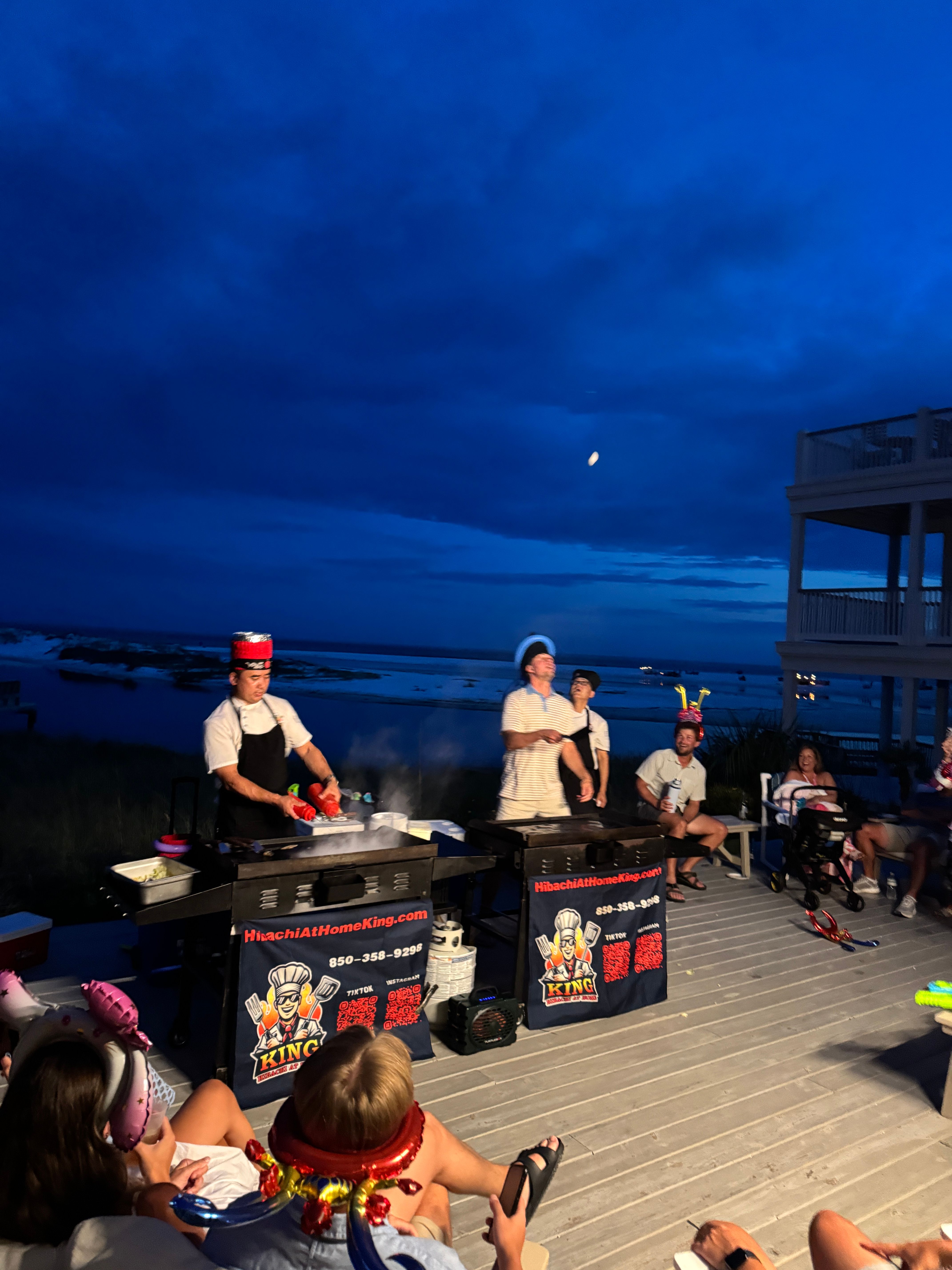 Evening beachfront hibachi show on a wooden deck — chefs cooking on portable griddles while a festive crowd in party hats watches beneath a deep blue sky and moon over the ocean.