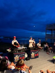 Evening beachfront hibachi show on a wooden deck — chefs cooking on portable griddles while a festive crowd in party hats watches beneath a deep blue sky and moon over the ocean.