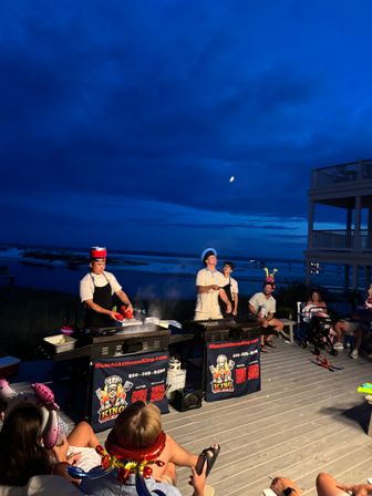 Evening beachfront hibachi show on a wooden deck — chefs cooking on portable griddles while a festive crowd in party hats watches beneath a deep blue sky and moon over the ocean.